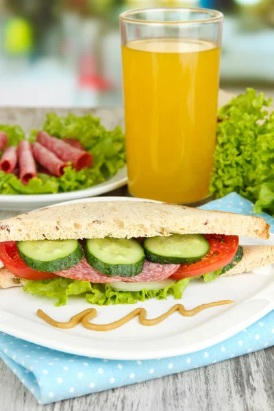 Composition with fruit juice and tasty sandwich with salami sausage and vegetables on color napkin, on wooden table background — Stock Photo, Image