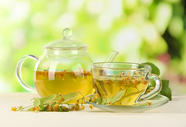 Kettle and cup of tea with linden on wooden table on nature background — Stock Photo, Image