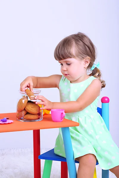 Schattig meisje zittend op weinig stoel in de buurt van tabel en smakelijke cookie, eten op grijze achtergrond — Stockfoto