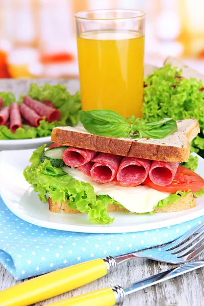 Composition with fruit juice and tasty sandwich with salami sausage and vegetables on color napkin, on wooden table background — Stock Photo, Image