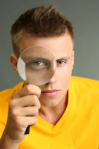 Young man looking through magnifying glass on grey background — Stock Photo, Image