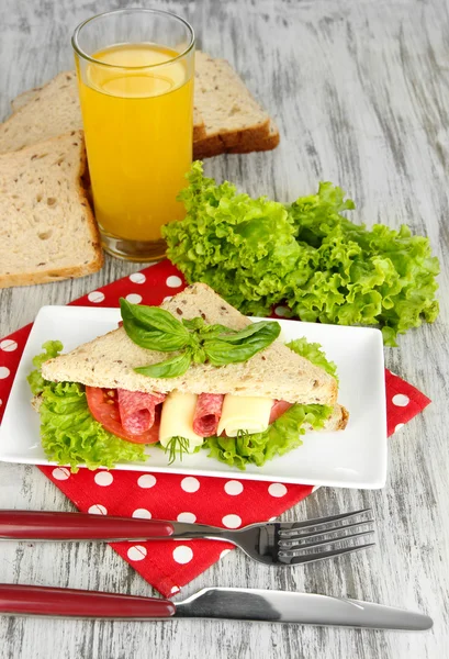Composición con zumo de fruta y sabroso sándwich con salchicha salami y verduras en servilleta de color, sobre fondo de mesa de madera — Foto de Stock