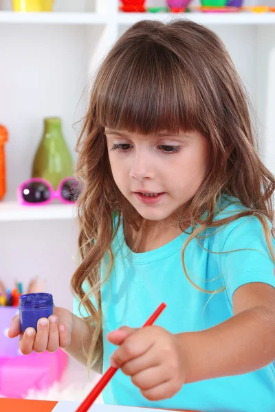 Niña dibuja sentado en la mesa en la habitación en los estantes de fondo —  Fotos de Stock