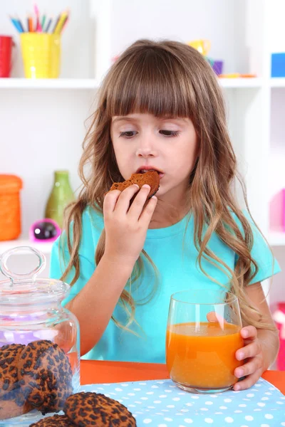 Little girl eating cookies and drinking juice sitting at table in room on shelves background — Stock Photo, Image