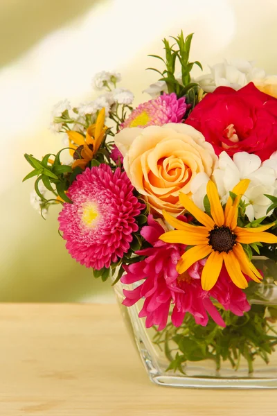 Beautiful bouquet of bright flowers in glass vase, on wooden table, on bright background — Stock Photo, Image