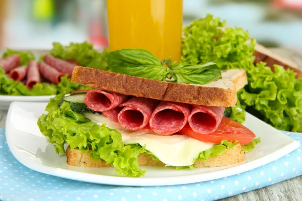 Composition with fruit juice and tasty sandwich with salami sausage and vegetables on color napkin, on wooden table background — Stock Photo, Image