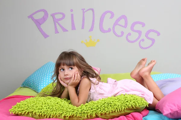 Little girl lying on bed in room on grey wall background — Stock Photo, Image