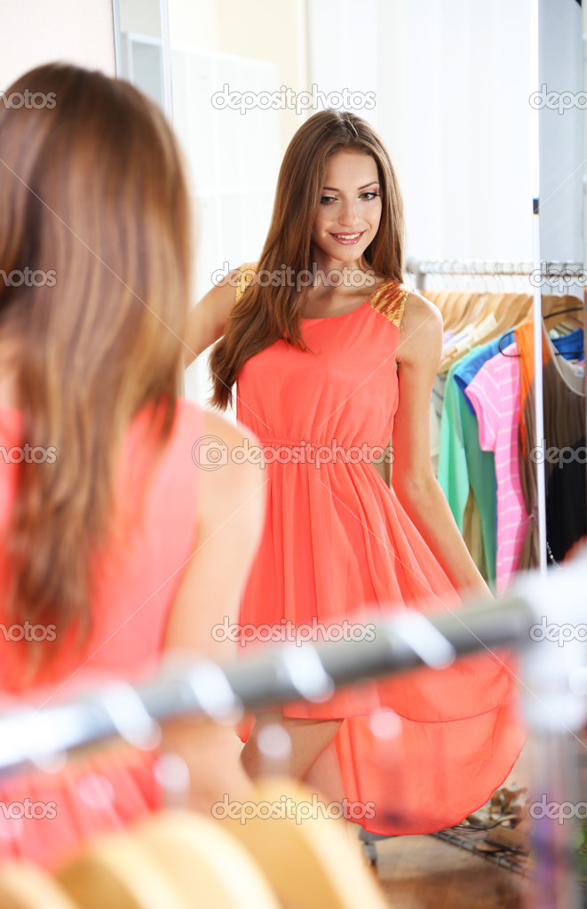 Beautiful girl trying dress near mirror on room background