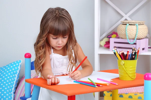 Маленькая девочка рисует сидя за столом в комнате на сером фоне стены — стоковое фото