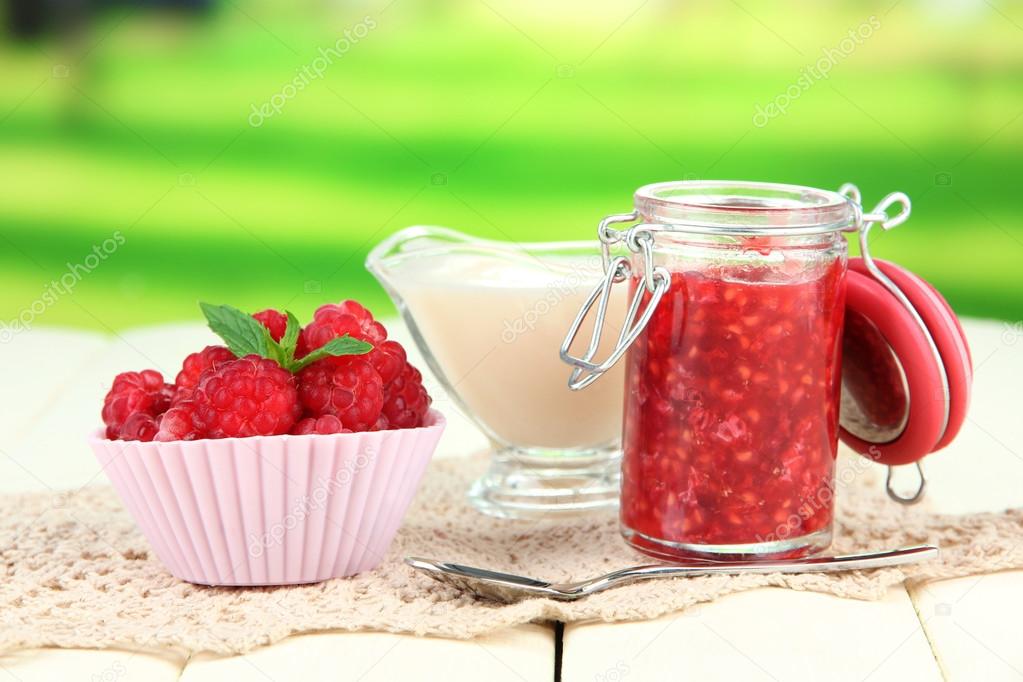 Fresh raspberry, raspberry jam and cream in jug on wooden table, on bright background