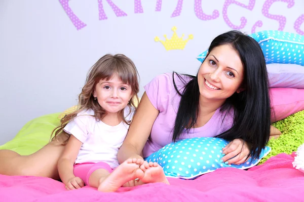 Маленька дівчинка з мамою лежить на ліжку в кімнаті на сірому фоні стіни — стокове фото