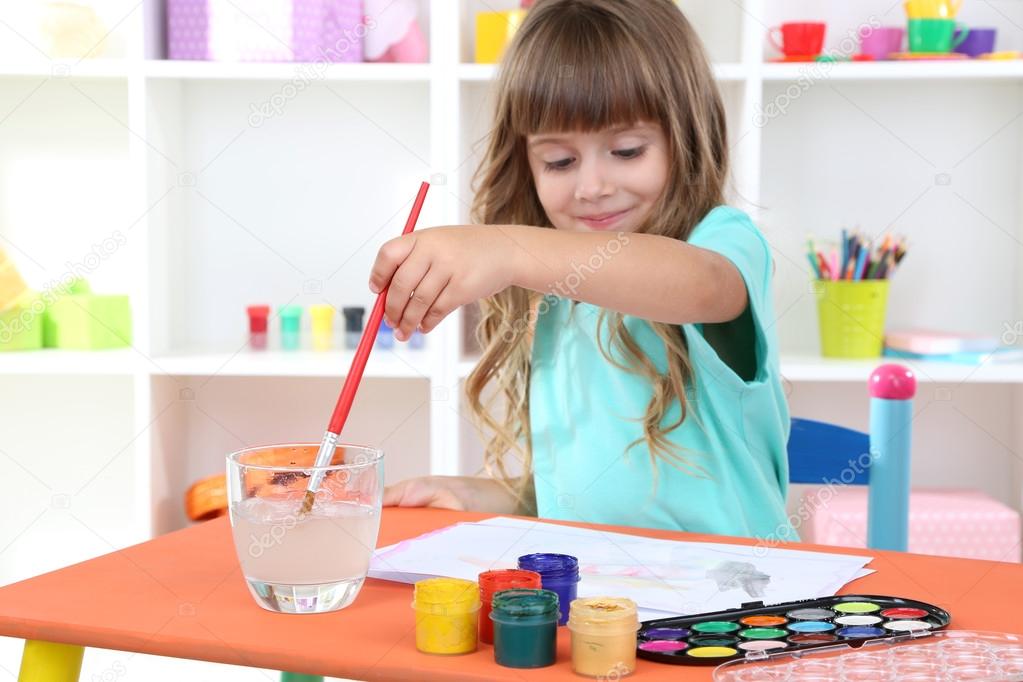 Little girl draws sitting at table in room on shelves background