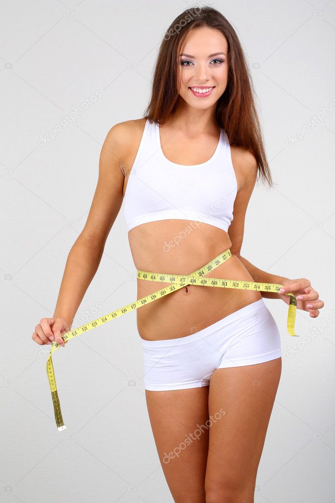 Beautiful young woman measuring her body with tape on grey background