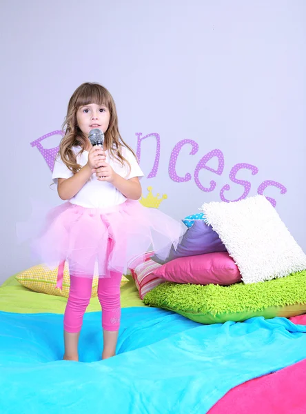 Little girl sings on bed in room on grey wall background — Stock Photo, Image