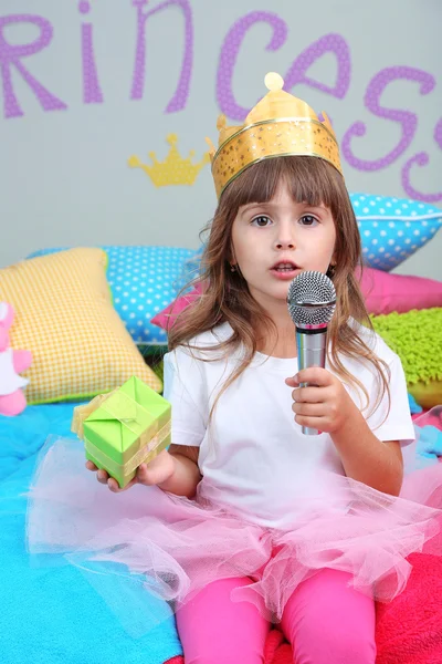 Маленька дівчинка сидить на ліжку з подарунком у кімнаті на сірому фоні стіни — стокове фото