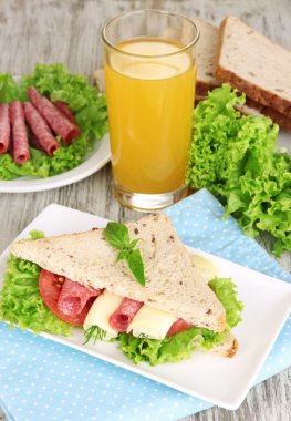 Lezzetli sandviç salam sosis ve ahşap masa arka plan üzerinde renkli peçete üzerinde sebze ve meyve suyu ile kompozisyon