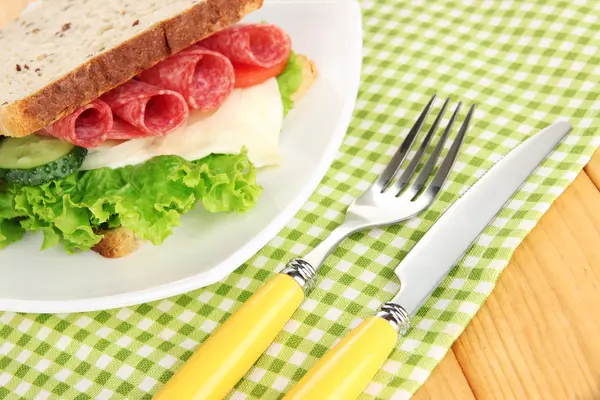 Tasty sandwich with salami sausage and vegetables on white plate, on wooden background — Stock Photo, Image