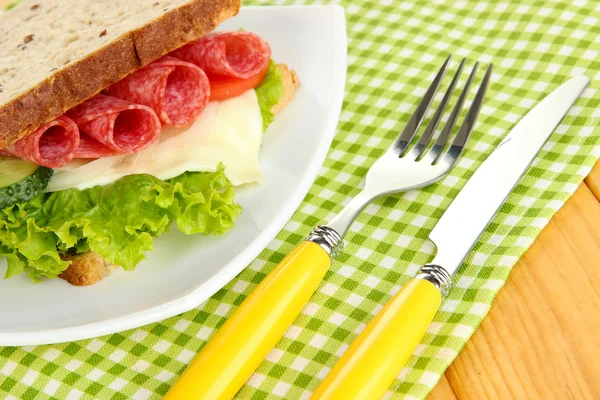 Tasty sandwich with salami sausage and vegetables on white plate, on wooden background — Stock Photo, Image