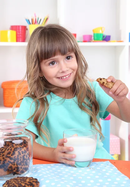 Meisje cookies eten en drinken van melk zittend aan tafel in kamer op planken achtergrond — Stockfoto