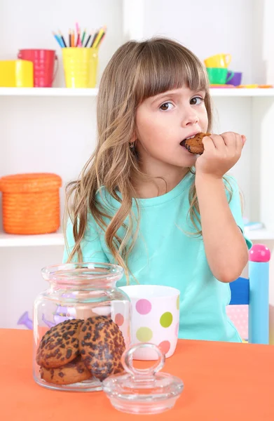 Menina comendo biscoitos sentados à mesa no quarto nas prateleiras fundo — Fotografia de Stock