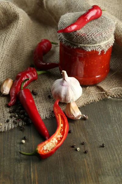 Composição com molho de salsa em jarra de vidro, pimenta vermelha e alho, sobre pano de saco, sobre fundo de madeira — Fotografia de Stock