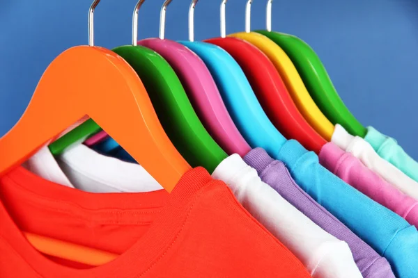 Verschillende shirts op kleurrijke hangers op blauwe achtergrond — Stockfoto