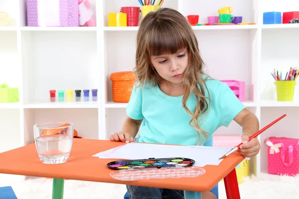 Menina desenha sentado à mesa no quarto em prateleiras fundo — Fotografia de Stock