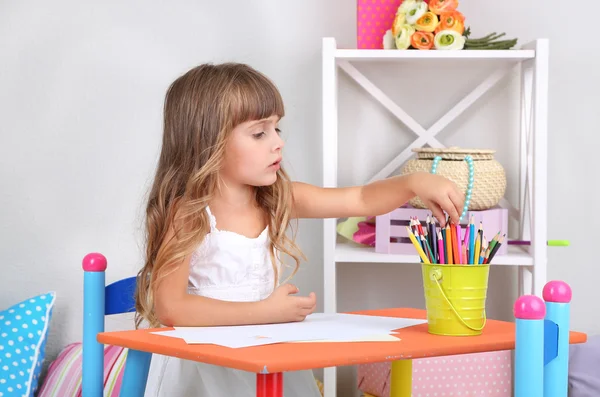 Niña dibuja sentado en la mesa en la habitación sobre fondo gris de la pared —  Fotos de Stock