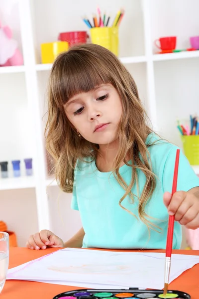 Niña dibuja sentado en la mesa en la habitación en los estantes de fondo — Foto de Stock