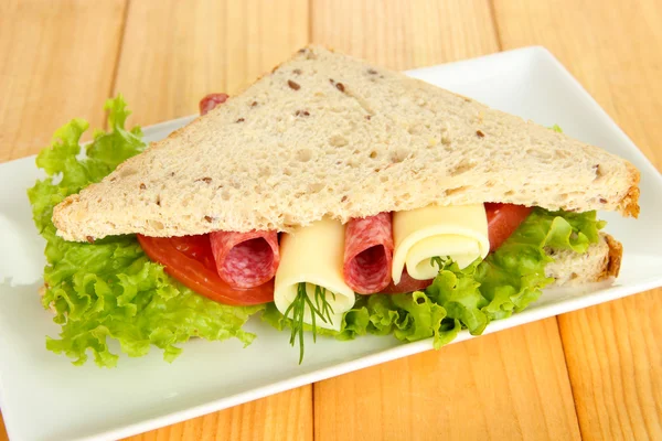 Tasty sandwich with salami sausage and vegetables on white plate, on wooden background — Stock Photo, Image