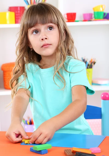 Weinig meisje mallen van plasticine zittend aan tafel in kamer op planken achtergrond — Stockfoto