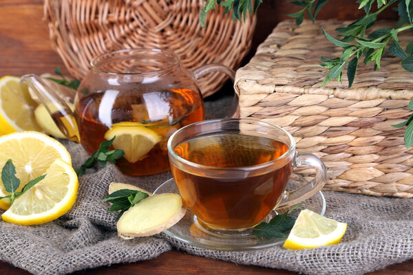 Чайник и чашка чая с имбирем на мешковине на деревянном столе на ярком фоне
