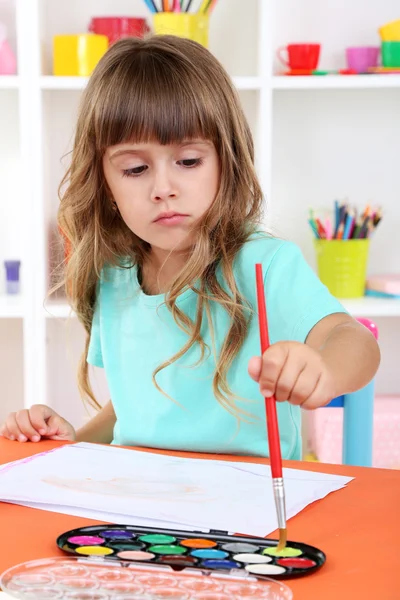 Niña dibuja sentado en la mesa en la habitación en los estantes de fondo — Foto de Stock