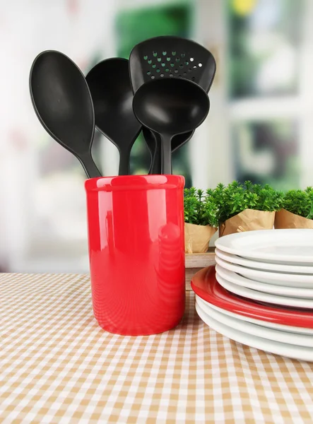 Plastic kitchen utensils in stand with clean dishes on tablecloth on bright background — Stock Photo, Image