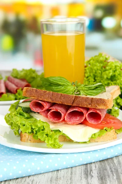 Composition with fruit juice and tasty sandwich with salami sausage and vegetables on color napkin, on wooden table background — Stock Photo, Image