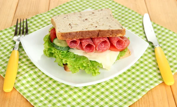 Вкусный сэндвич с колбасой и овощами на белой тарелке, на деревянном фоне — стоковое фото