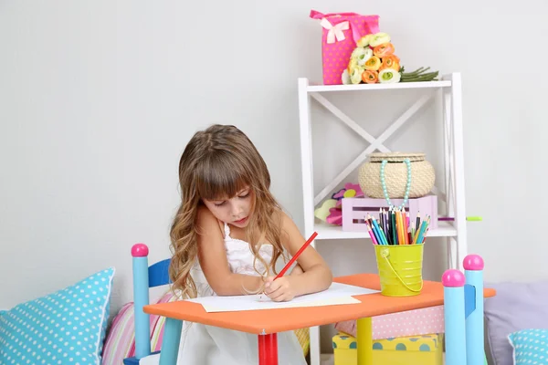 Niña dibuja sentado en la mesa en la habitación sobre fondo gris de la pared —  Fotos de Stock