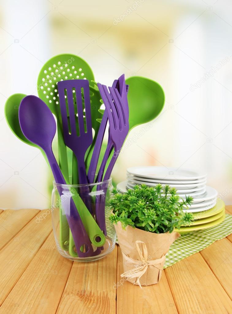 Plastic kitchen utensils in stand with clean dishes on table on bright background