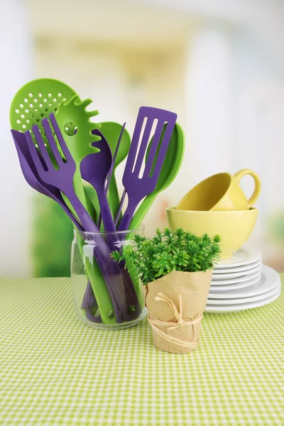 Utensílios de cozinha de plástico em stand com pratos limpos na toalha de mesa em fundo brilhante — Fotografia de Stock