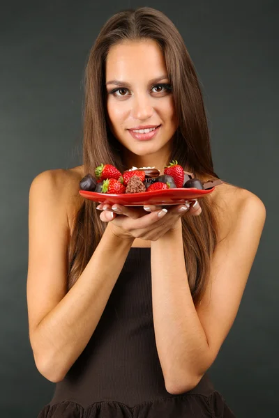 Retrato de una hermosa joven con dulces de chocolate en el plato sobre fondo gris — Foto de Stock