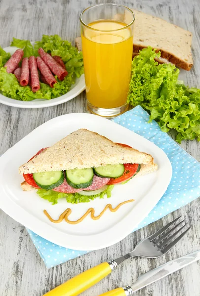 Composizione con succo di frutta e gustoso sandwich con salsiccia di salame e verdure su tovagliolo di colore, su sfondo tavolo di legno — Foto Stock