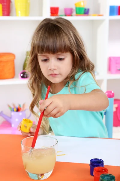 Маленькая девочка рисует сидя за столом в комнате на фоне полок — стоковое фото