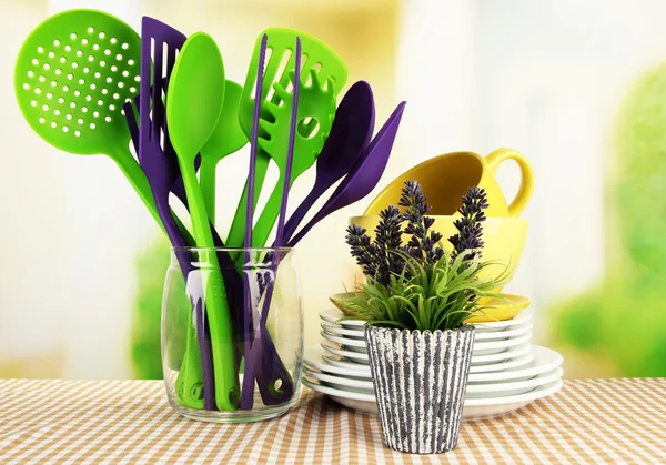 Plastic kitchen utensils in stand with clean dishes on tablecloth on bright background — Stock Photo, Image