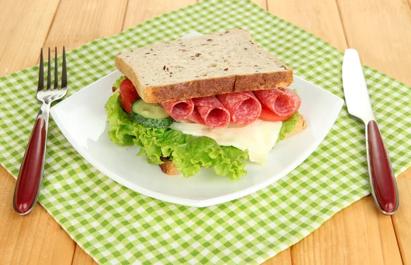 Tasty sandwich with salami sausage and vegetables on white plate, on wooden background — Stock Photo, Image