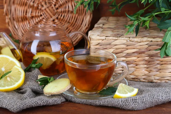 Hervidor de agua y taza de té con jengibre sobre tela de saco sobre mesa de madera sobre fondo brillante —  Fotos de Stock