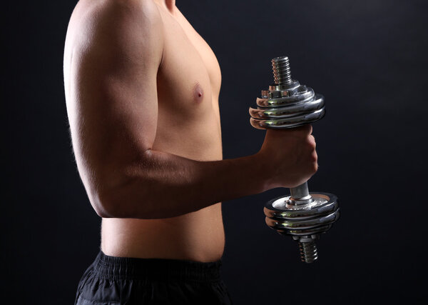 Handsome young muscular sportsman execute exercise with dumbbell on dark background