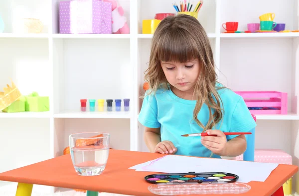 Маленька дівчинка малює, сидячи за столом у кімнаті на фоні полиць — стокове фото