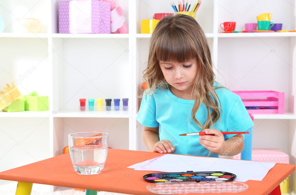 Little girl draws sitting at table in room on shelves background