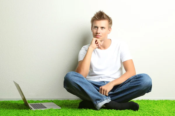 Hombre joven que se relaja en la alfombra con el ordenador portátil, sobre fondo de pared gris — Foto de Stock