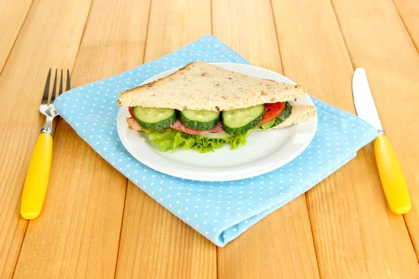 Tasty sandwich with salami sausage and vegetables on white plate, on wooden background — Stock Photo, Image